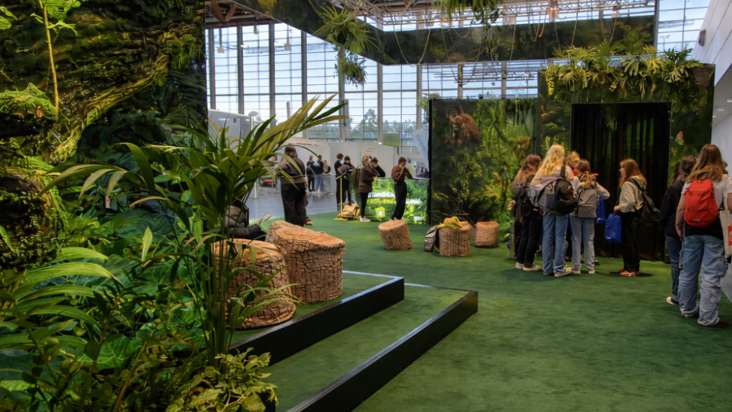 Messestand / Event / Berufsorientierung - Messestand für BOBY – Berufsorientierung Bayern Überblick über den begrünten BOBY-Messestand mit Farnwänden, Baumstumpf-Sitzgelegenheiten und Besuchergruppen in der Messehalle