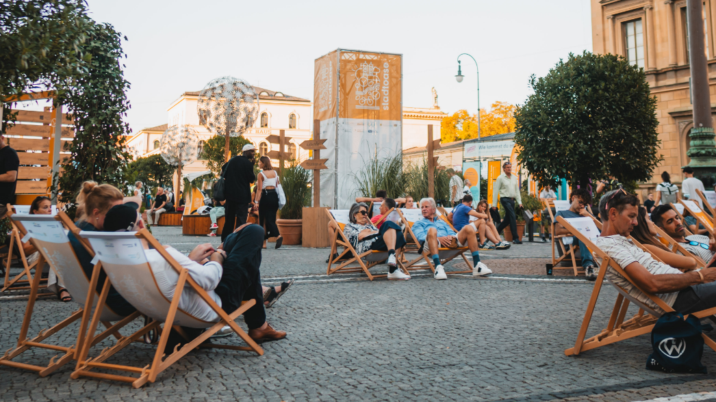Event / öffentlicher Raum / Klimakommunikation - Raum für Klimaschutz und Mobilität Weitwinkelansicht der Stadtoase mit Liegestühlen, Pusteblumen-Skulpturen, Infosäule und entspannten Besucher:innen im Abendlicht
